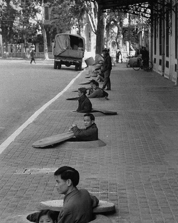 Des habitants d’Hanoi attendent dans des abris de trottoir qui leur descendent jusqu’à la poitrine le signal de fin d’alerte, pendant une alerte au raid aérien. Hanoi, Nord-Vietnam, 1967.