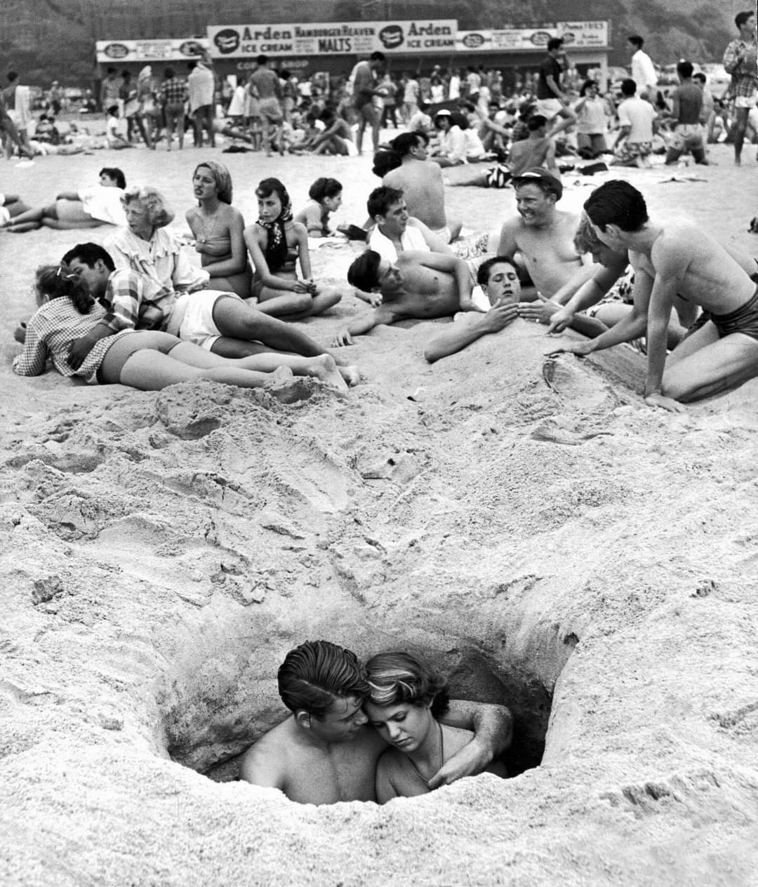 Un couple se fait des câlins en s’asseyant dans un trou alors que d’autres profitent de la plage le 4 juillet à Santa Monica, Californie, 1950. (Photographié par Ralph Crane)