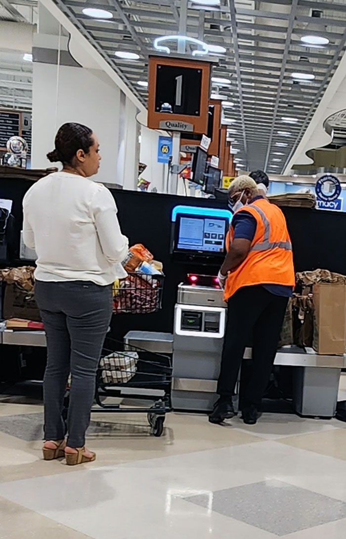 Une femme se rend à la caisse automatique, demande au préposé de décharger son chariot, de scanner et d’emballer chaque article.